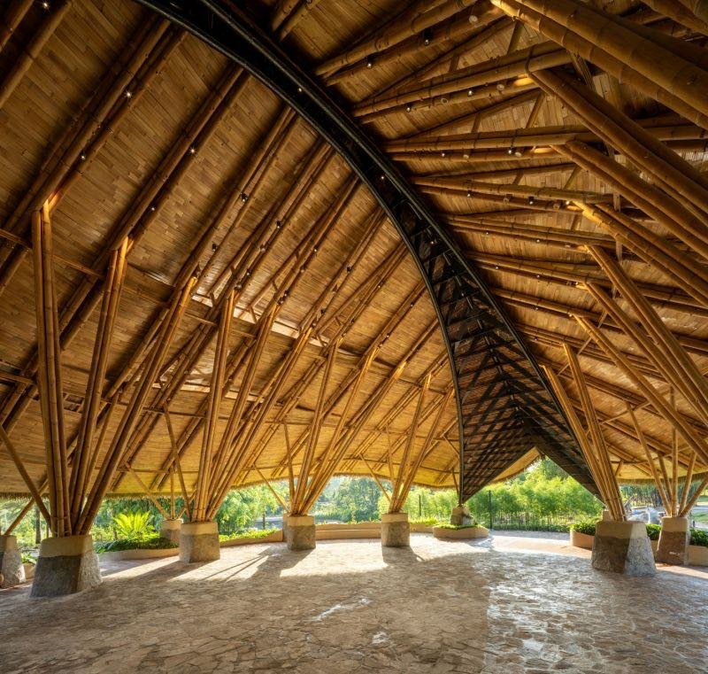 Bamboo roof on a steel arch over an open stone floor at Kaway'an Eco Park, a curved bamboo walkway, and the pavilion lit at dusk